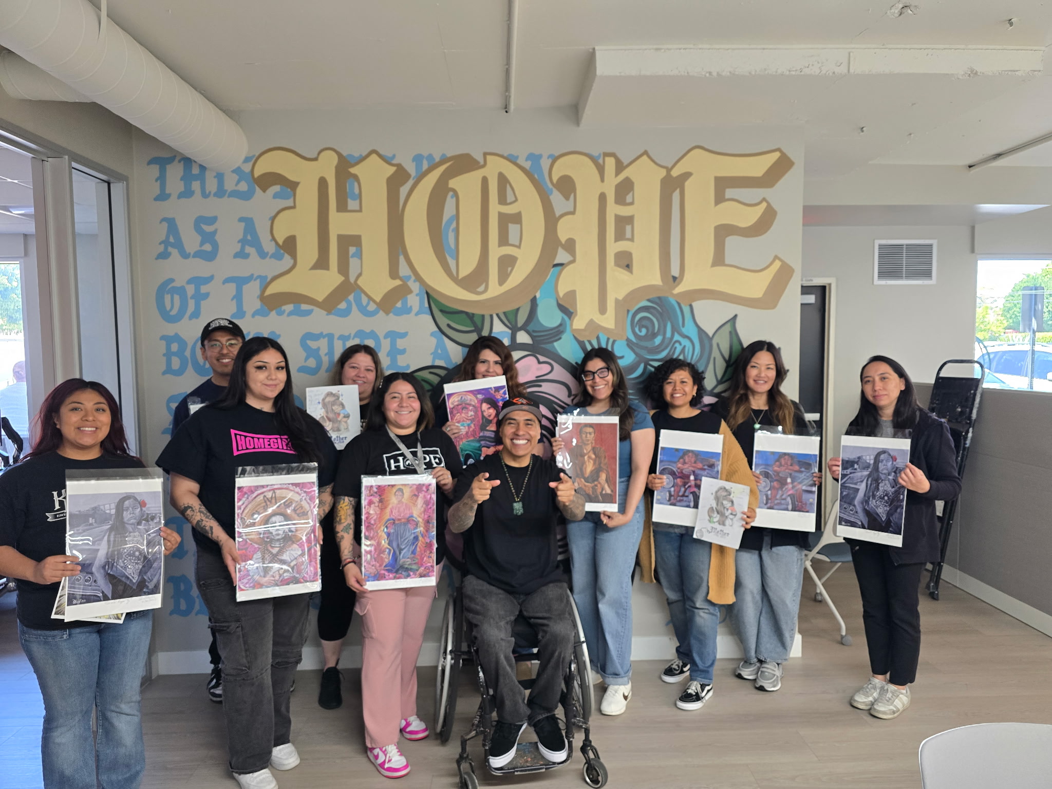 Diverse group of people, including one in a wheelchair, smiling and holding up various artworks in front of a "HOPE" mural.