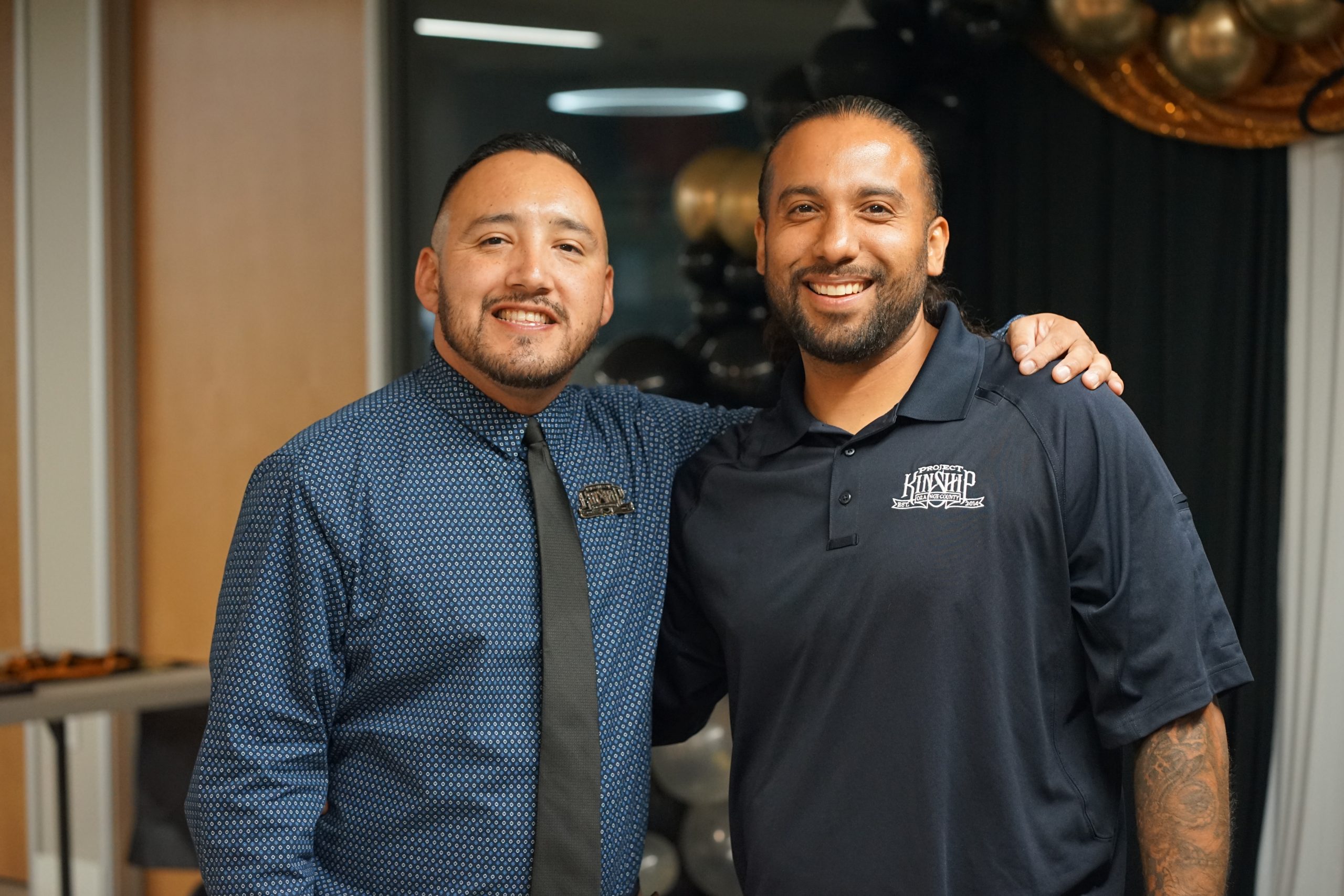 Two smiling men, one in a blue patterned shirt and tie, the other in a black polo with a 'Project Kinship' logo, stand toget…