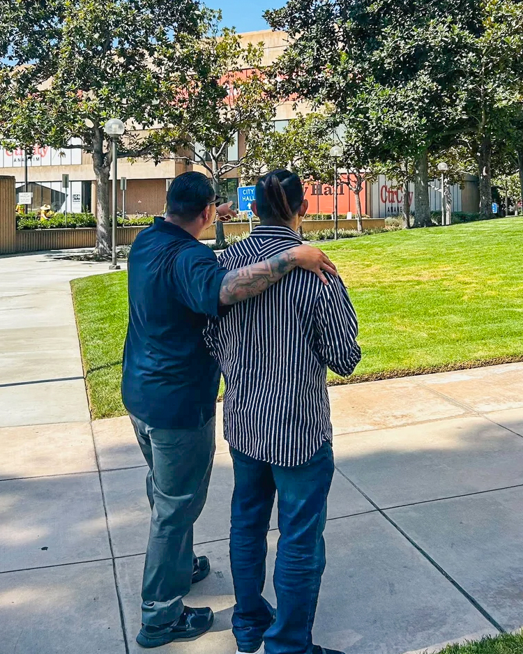 Two men from behind, one with arm around the other, pointing towards Anaheim City Hall. Sunny outdoor scene with trees and g…