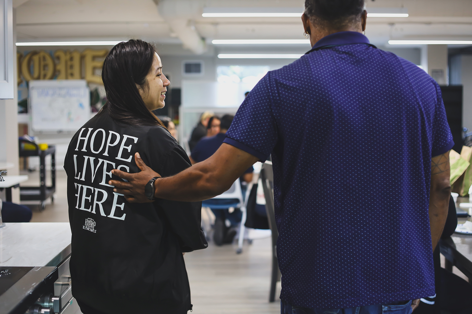 A man in a blue polka-dotted shirt places his hand on the back of a smiling woman wearing a black shirt that reads "HOPE LIV…