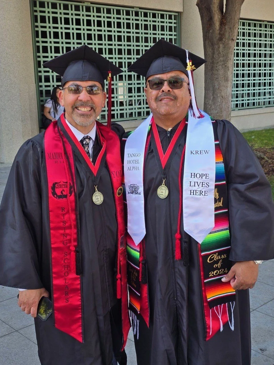 Proud Graduates Celebrate Academic Success in Regalia Two smiling men in black graduation gowns, caps, and stoles, wearing sunglasses, celebrating their academic achievement.