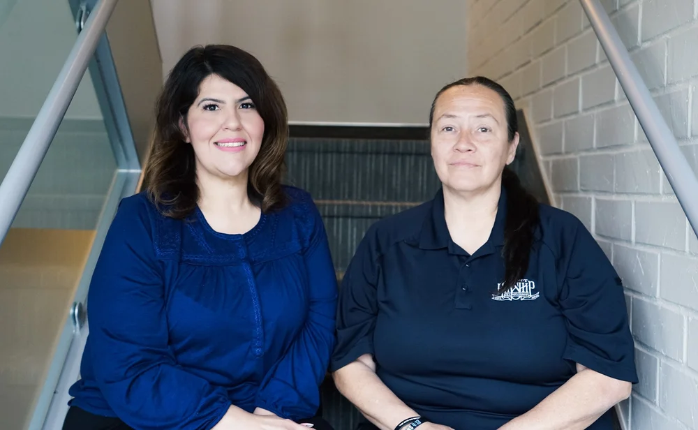 Two women, one with dark wavy hair and a blue blouse, the other with dark hair in a ponytail and a navy polo, sit on stairs.