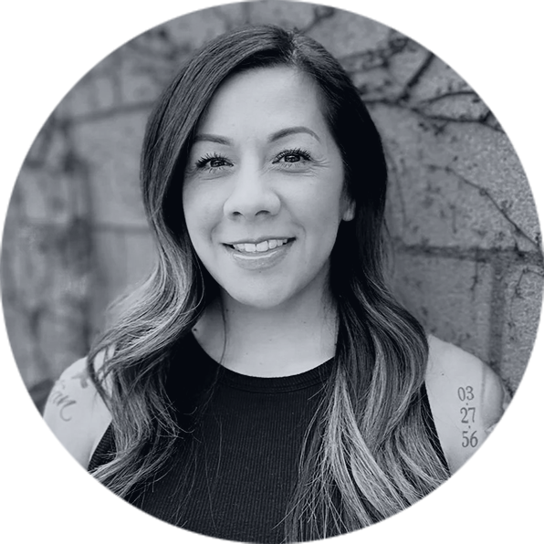 Smiling Professional Woman Headshot for Executive Team Confident smiling woman with long wavy hair and arm tattoos, wearing a dark tank top, against a stone wall.