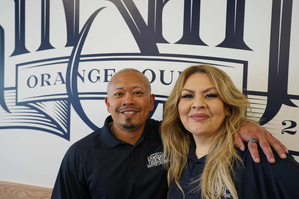 Man with goatee and woman with long blonde hair smiling, wearing Project Kinship polo shirts, in front of an "ORANGE COUNTY"…
