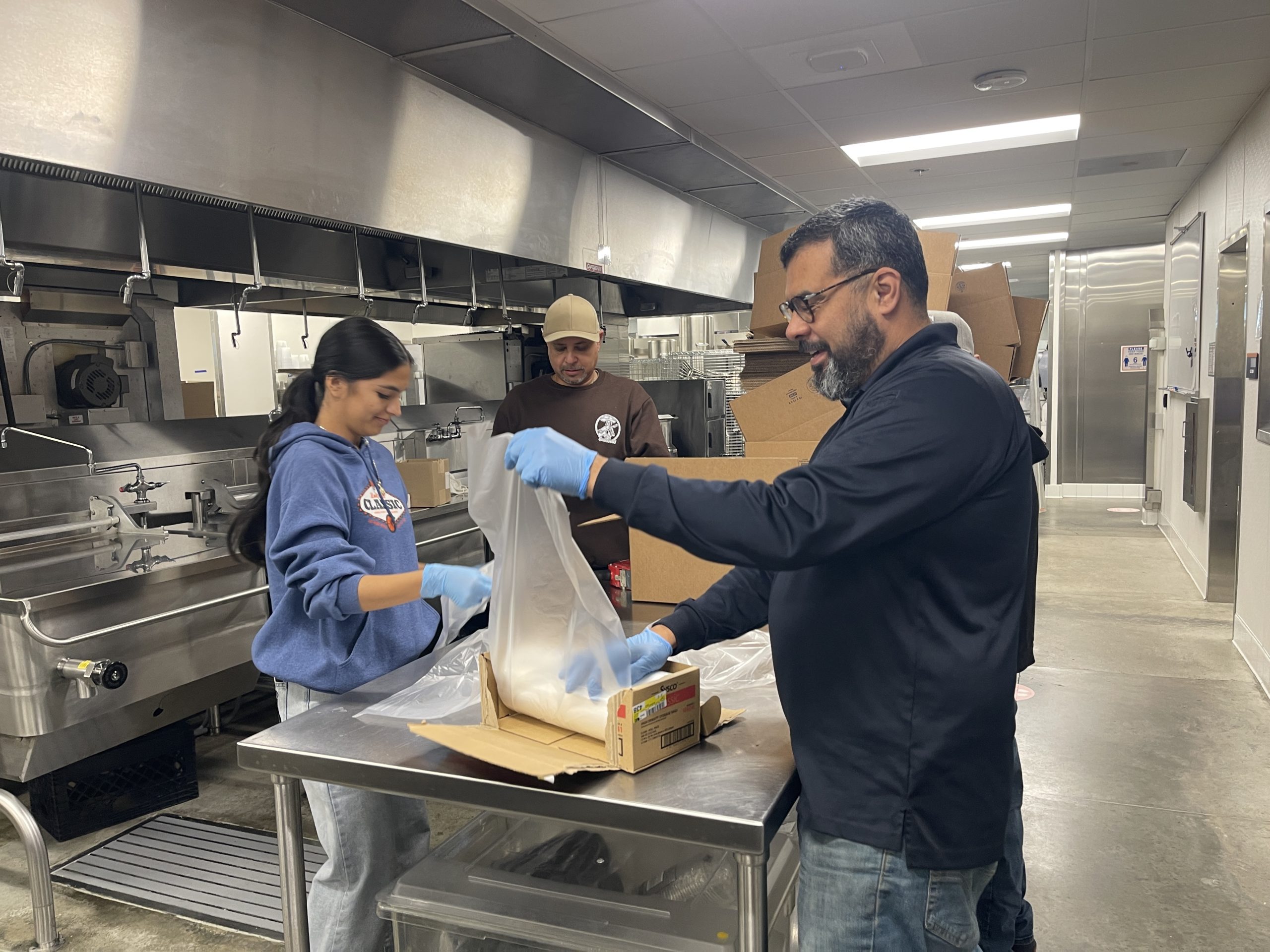 Workforce Team Packaging Food Supplies in Commercial Kitchen Three team members in a commercial kitchen, wearing gloves, efficiently package bulk ingredients into a box on a stainless s…