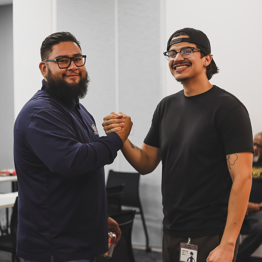 Two men, one bearded in glasses and a navy jacket, the other in a black t-shirt and backward cap, shake hands and smile.