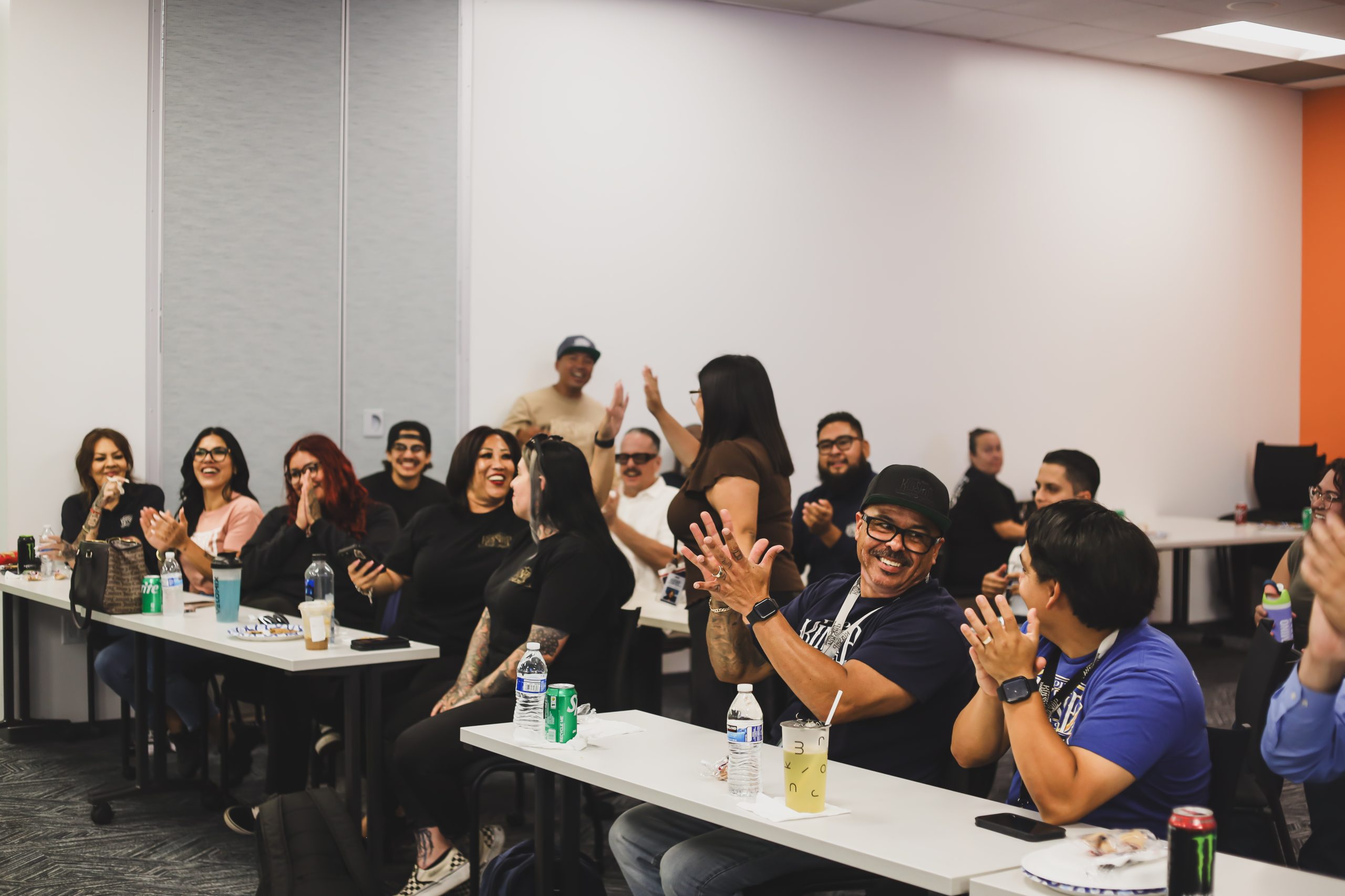 Diverse group of people in a classroom setting, many smiling and clapping, with drinks and snacks on tables.