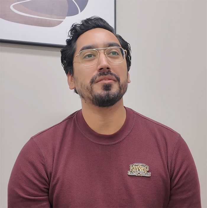 Man with beard and gold-rimmed glasses looking up, wearing a maroon waffle-knit shirt with a 'PROJECT KINSHIP' pin.