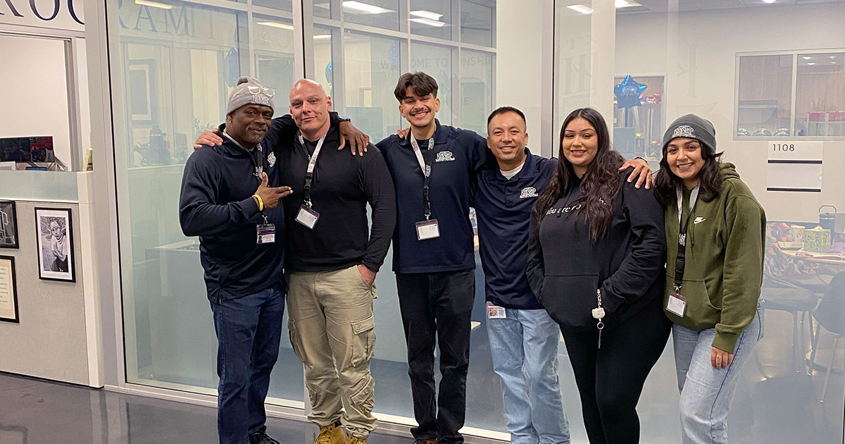 Six diverse colleagues, four men and two women, smiling and standing together in a modern office environment.
