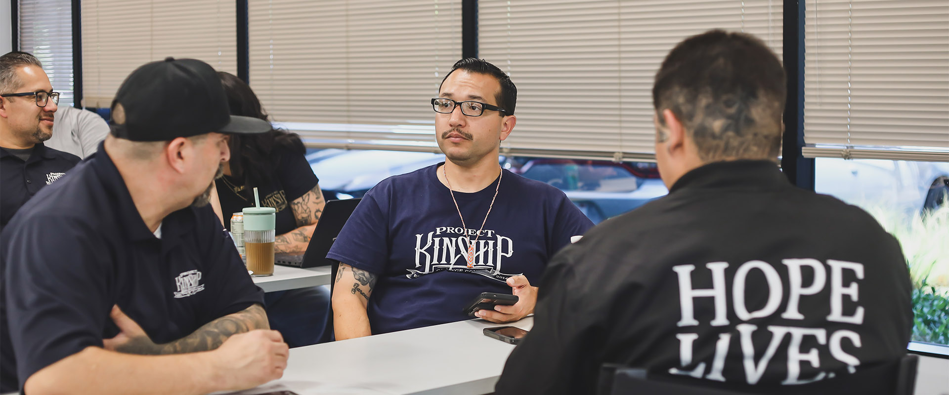 Men in Project Kinship shirts attending a meeting, one with 'HOPE LIVES' on his back, discussing in a bright room.