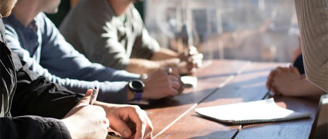 Four men at a wooden table, one actively writing notes on a yellow pad during a focused group discussion.