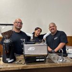Professional Barista Team Ready for Coffee Service Three smiling baristas, two men and one woman, stand behind a mobile coffee bar with a grinder, espresso machine, and sink.