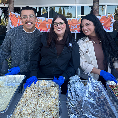 Volunteers Serve Thanksgiving Meal with Smiles Three volunteers, two women and one man, smile while serving Thanksgiving food from large catering trays, wearing blue glove…