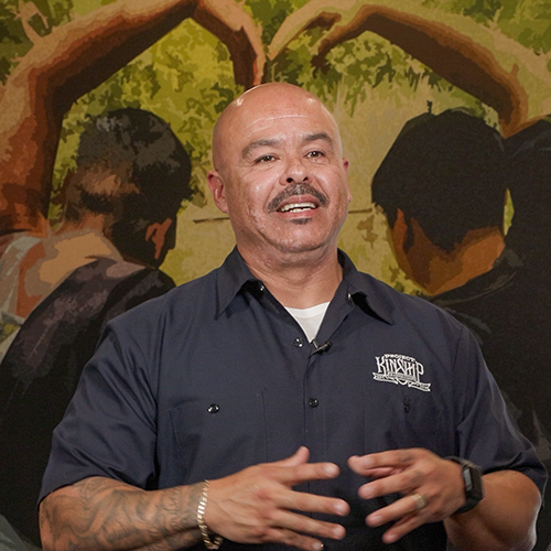 Man in Kinship Shirt Speaking Against Mural Background Bald man with mustache and goatee, wearing a dark blue work shirt with 'KINSHIP' logo, gesturing while speaking, in front of…