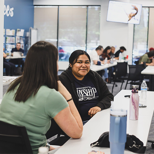 Project Kinship Participants Engaging in Community Discussion Two women sit at a table, one smiling and wearing a 'Project Kinship' hoodie, in a bright, modern community space.