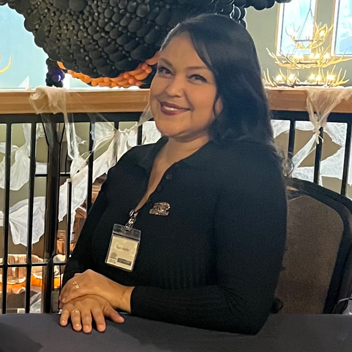 April, a smiling woman with dark hair and a black collared shirt, wearing an ID badge, sits at a table with hands clasped.