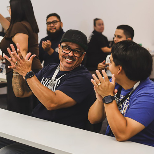 Engaged Participants Clap at Kinship Learns Event Two men, one in a black cap and glasses, smiling and clapping enthusiastically at a Kinship Learns event. Others clap nearby.