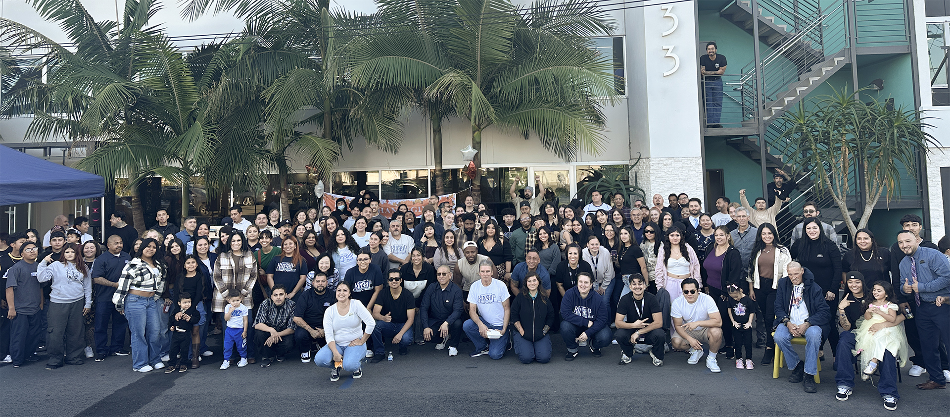 A large, diverse group of people, including adults and children, gathered outdoors in front of a modern building with palm t…