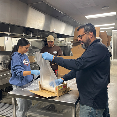 Workforce Program Participants Packaging Food Three individuals in a commercial kitchen, wearing gloves, collaboratively packaging white ingredients into cardboard boxes…