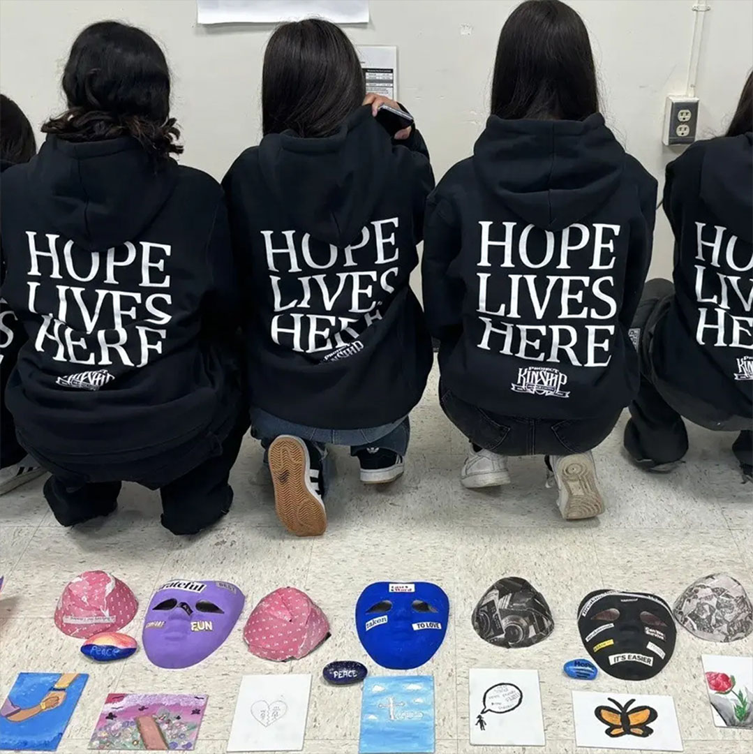Hope Lives Here: Youth Art and Expression Three people in black 'HOPE LIVES HERE' hoodies crouch, viewing colorful masks, painted rocks, and small artworks on the flo…
