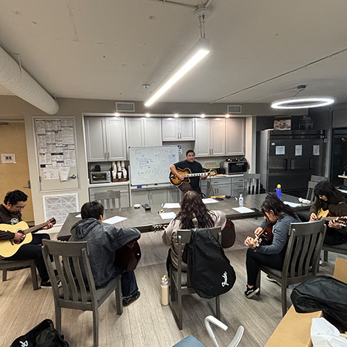 Group Guitar Lessons at the Healing Institute An instructor plays guitar for five students practicing acoustic guitars around a large table in a modern common room.