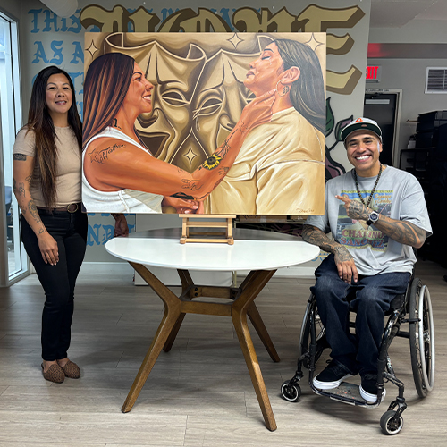 Artists Present Inspiring Painting at Healing Institute Artists, a standing woman and a man in a wheelchair, proudly showcase their painting of two women, theatrical masks, and cel…